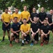 A group of approximately 11 military personnel pose for a formal group photo on a grass field. The group includes members from both the U.S. Navy (wearing gold/yellow 'NAVY' shirts and navy blue shorts) and U.S. Army (wearing black shirts and camouflage uniform). A senior Naval officer in dress whites stands on the far left, and an Army soldier in full ACU/OCP camouflage with a distinctive hat stands on the far right.