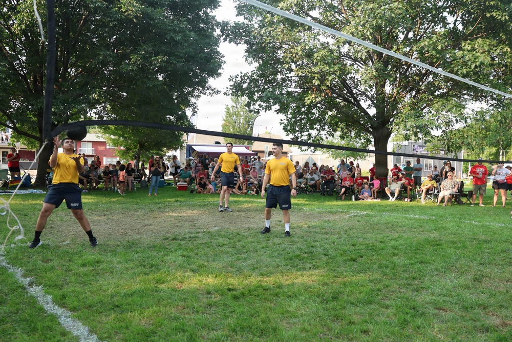 Three individuals wearing matching yellow shirts and dark shorts with 'NAVY' branding are playing volleyball on a grass court outdoors. A large crowd of spectators is gathered in the background watching the game. The setting appears to be a community or public event held in a park-like area.