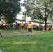 Three individuals wearing matching yellow shirts and dark shorts with 'NAVY' branding are playing volleyball on a grass court outdoors. A large crowd of spectators is gathered in the background watching the game. The setting appears to be a community or public event held in a park-like area.