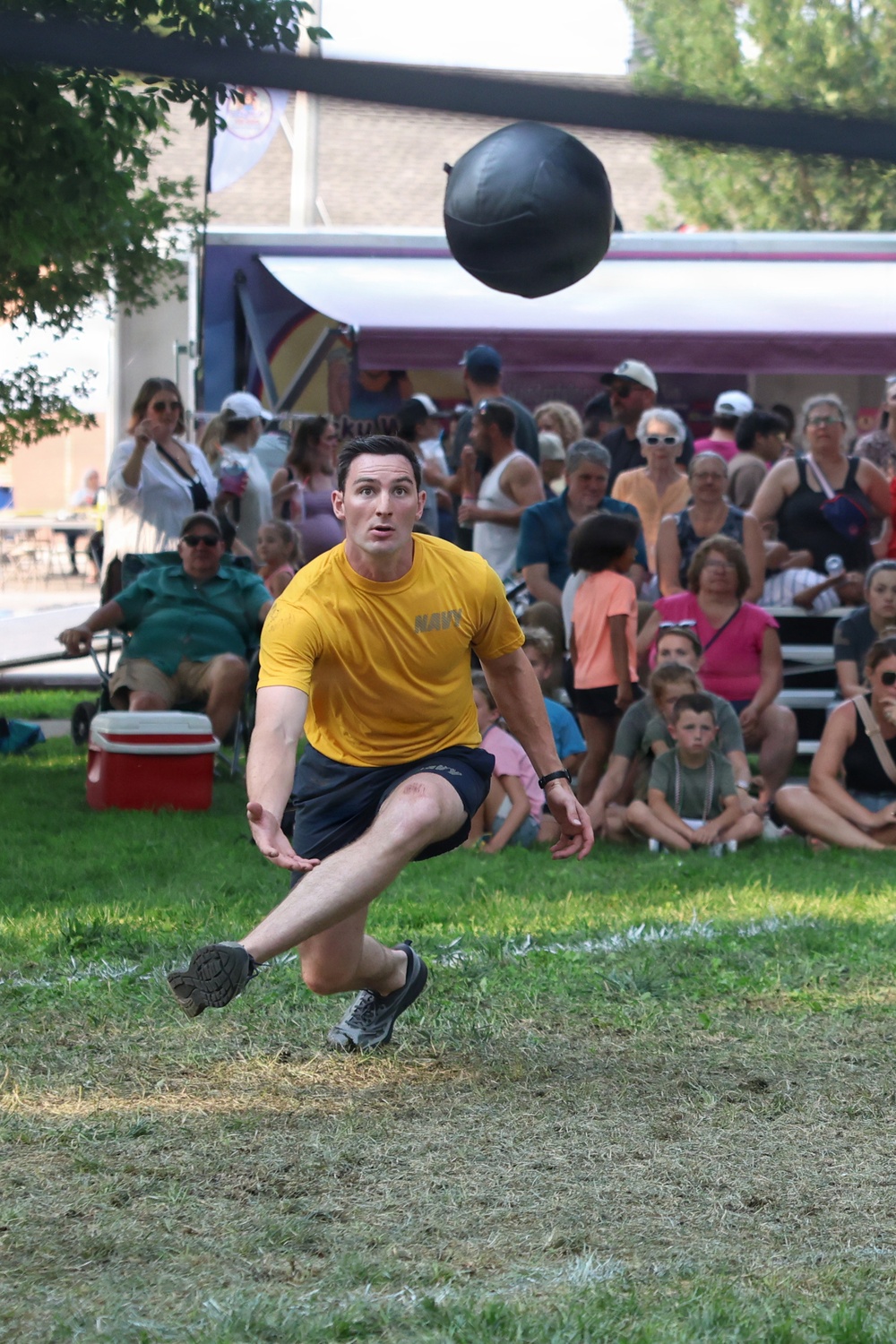 A man wearing a yellow 'NAVY' t-shirt and dark shorts is captured mid-action on a grass field, appearing to have just thrown or released a large dark medicine ball or shot put-style ball into the air. He is in a low, lunging stance with both feet near the ground, suggesting a powerful release. A large crowd of spectators is gathered in the background, watching the event.