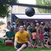 A man wearing a yellow 'NAVY' t-shirt and dark shorts is captured mid-action on a grass field, appearing to have just thrown or released a large dark medicine ball or shot put-style ball into the air. He is in a low, lunging stance with both feet near the ground, suggesting a powerful release. A large crowd of spectators is gathered in the background, watching the event.