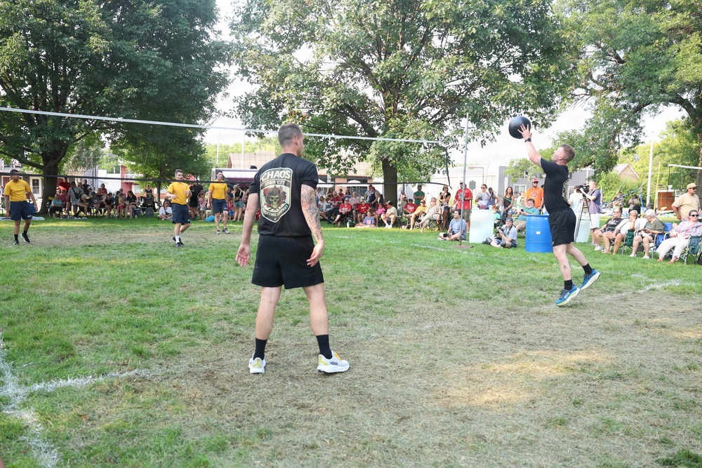 An outdoor volleyball game is taking place on a grass court in what appears to be a public park during a community event. Two teams are visible, with one team wearing black shirts and the other wearing yellow shirts. A large crowd of spectators is seated and standing along the sidelines watching the match.