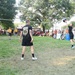 An outdoor volleyball game is taking place on a grass court in what appears to be a public park during a community event. Two teams are visible, with one team wearing black shirts and the other wearing yellow shirts. A large crowd of spectators is seated and standing along the sidelines watching the match.