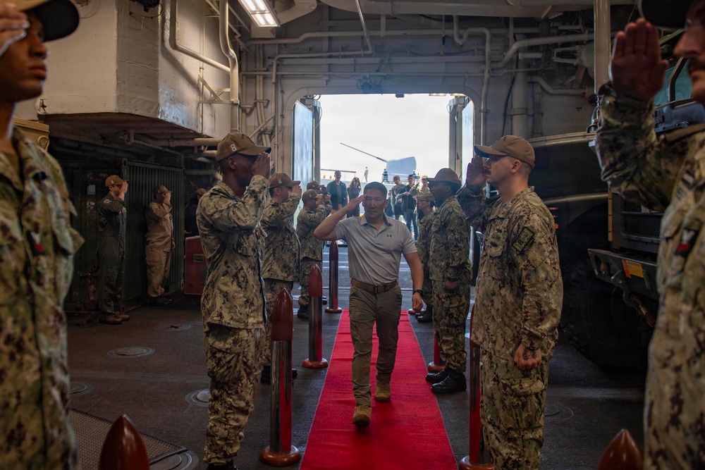 A civilian or dignitary in civilian attire (white polo shirt, khaki pants) walks along a red carpet through a formal military sideboy honors ceremony aboard what appears to be a U.S. Navy vessel. Uniformed sailors in Navy Working Uniform (NWU) Type III camouflage are lined up on both sides rendering salutes. The setting appears to be the well deck or hangar bay of a naval ship, indicated by the large open stern gate and industrial interior.