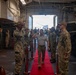 A civilian or dignitary in civilian attire (white polo shirt, khaki pants) walks along a red carpet through a formal military sideboy honors ceremony aboard what appears to be a U.S. Navy vessel. Uniformed sailors in Navy Working Uniform (NWU) Type III camouflage are lined up on both sides rendering salutes. The setting appears to be the well deck or hangar bay of a naval ship, indicated by the large open stern gate and industrial interior.