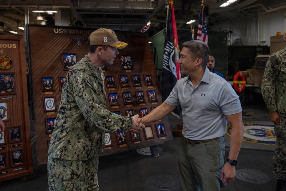A U.S. Navy sailor in digital camouflage uniform and cover is shaking hands with a civilian man wearing a gray polo shirt aboard what appears to be the USS Makin Island (LHD-8), as indicated by the signage visible in the background. The setting appears to be the ship's hangar bay or interior deck area, decorated with command plaques, portrait boards, and military flags including a POW/MIA flag and an American flag. The interaction appears to be a formal greeting or recognition ceremony.