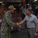A U.S. Navy sailor in digital camouflage uniform and cover is shaking hands with a civilian man wearing a gray polo shirt aboard what appears to be the USS Makin Island (LHD-8), as indicated by the signage visible in the background. The setting appears to be the ship's hangar bay or interior deck area, decorated with command plaques, portrait boards, and military flags including a POW/MIA flag and an American flag. The interaction appears to be a formal greeting or recognition ceremony.