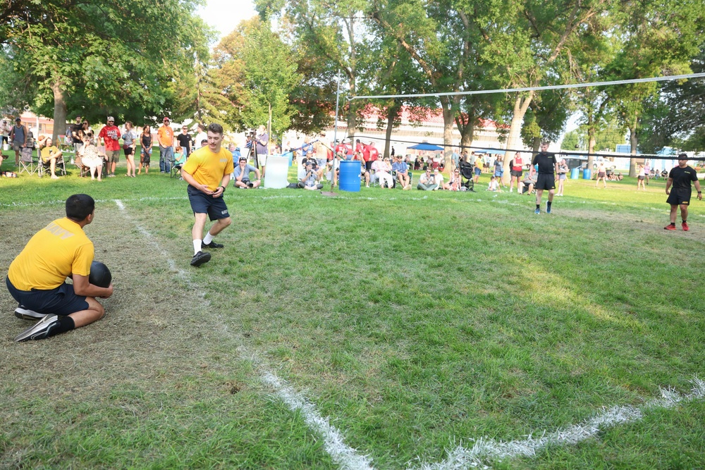 An outdoor sports event taking place on a grass field, likely a rugby or football game, with players in yellow and black uniforms actively participating. One player in yellow is running while another crouches near the sideline, and officials in black are visible on the field. A crowd of spectators lines the perimeter under large shade trees on a sunny day.