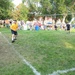 An outdoor sports event taking place on a grass field, likely a rugby or football game, with players in yellow and black uniforms actively participating. One player in yellow is running while another crouches near the sideline, and officials in black are visible on the field. A crowd of spectators lines the perimeter under large shade trees on a sunny day.