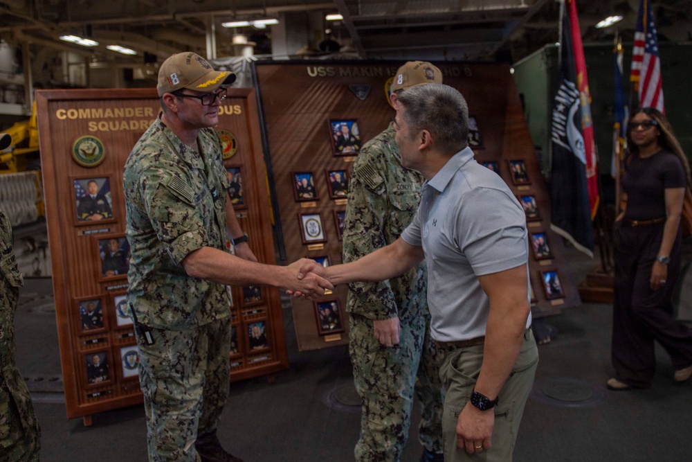A formal handshake is taking place aboard the USS Makin Island between a U.S. Navy sailor in camouflage uniform and a civilian or off-duty individual in a polo shirt and khaki pants. The setting appears to be the ship's hangar bay or interior deck, with command display boards visible in the background showing officer portraits. Additional military personnel and flags, including the U.S. flag and POW/MIA flag, are visible in the background.