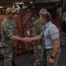 A formal handshake is taking place aboard the USS Makin Island between a U.S. Navy sailor in camouflage uniform and a civilian or off-duty individual in a polo shirt and khaki pants. The setting appears to be the ship's hangar bay or interior deck, with command display boards visible in the background showing officer portraits. Additional military personnel and flags, including the U.S. flag and POW/MIA flag, are visible in the background.