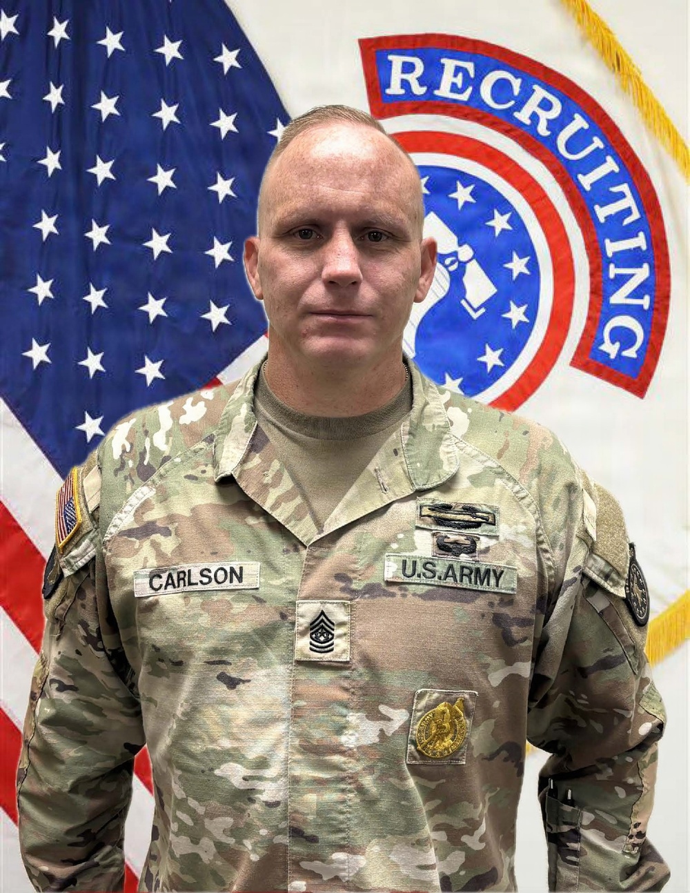A U.S. Army soldier named Carlson poses for an official portrait in Army Combat Uniform (ACU/OCP pattern). He stands in front of a U.S. flag and a U.S. Army Recruiting Command flag. His rank insignia indicates he holds the grade of Sergeant Major or Command Sergeant Major.