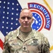 A U.S. Army soldier named Carlson poses for an official portrait in Army Combat Uniform (ACU/OCP pattern). He stands in front of a U.S. flag and a U.S. Army Recruiting Command flag. His rank insignia indicates he holds the grade of Sergeant Major or Command Sergeant Major.