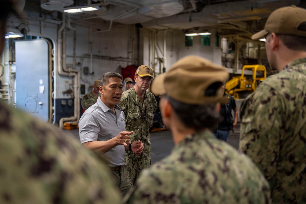A civilian in a gray polo shirt is speaking to a group of U.S. Navy personnel in digital camouflage uniforms aboard what appears to be a naval vessel. The setting is an interior deck or hangar bay area with visible pipes, equipment, and industrial infrastructure. The civilian appears to be briefing or addressing the assembled sailors.