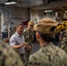 A civilian in a gray polo shirt is speaking to a group of U.S. Navy personnel in digital camouflage uniforms aboard what appears to be a naval vessel. The setting is an interior deck or hangar bay area with visible pipes, equipment, and industrial infrastructure. The civilian appears to be briefing or addressing the assembled sailors.