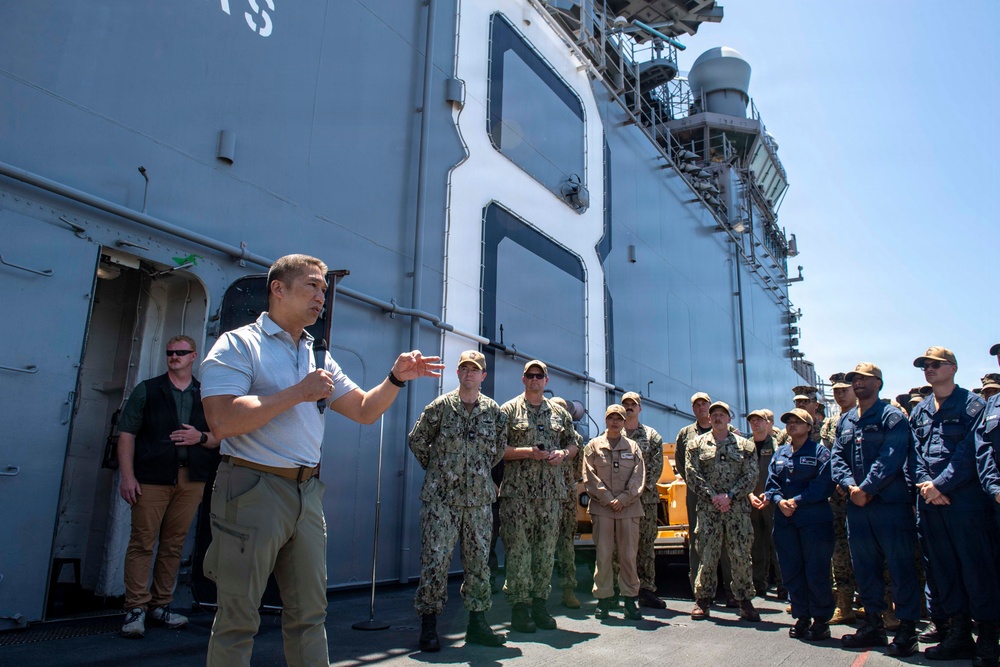 A civilian in a white polo shirt and khaki pants addresses a group of uniformed U.S. Navy personnel on the deck or pier alongside a large naval vessel. The gathering appears to be a formal briefing or address, with sailors standing attentively in two distinct uniform types: Navy Working Uniform (NWU) digital camouflage and tan/khaki uniforms. The large gray hull of a naval ship dominates the background.