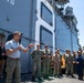 A civilian in a white polo shirt and khaki pants addresses a group of uniformed U.S. Navy personnel on the deck or pier alongside a large naval vessel. The gathering appears to be a formal briefing or address, with sailors standing attentively in two distinct uniform types: Navy Working Uniform (NWU) digital camouflage and tan/khaki uniforms. The large gray hull of a naval ship dominates the background.