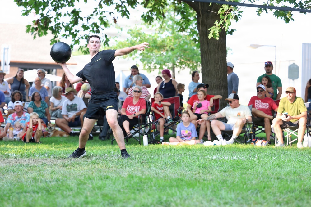 A man wearing a black Army uniform is preparing to throw a heavy black ball, likely a medicine ball or shot put, on a grass field during what appears to be a public outdoor event or competition. A large crowd of spectators is seated in lawn chairs and standing in the background, watching the activity. The event appears to be a community or military fitness demonstration held outdoors on a sunny day.