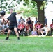 A man wearing a black Army uniform is preparing to throw a heavy black ball, likely a medicine ball or shot put, on a grass field during what appears to be a public outdoor event or competition. A large crowd of spectators is seated in lawn chairs and standing in the background, watching the activity. The event appears to be a community or military fitness demonstration held outdoors on a sunny day.