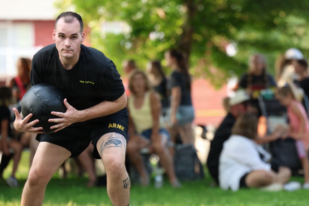 A U.S. Army soldier wearing black PT (physical training) uniform with 'ARMY' printed on the shorts is holding a medicine ball during what appears to be a fitness event or competition. The soldier has visible tattoos on his leg and is in a crouched athletic stance. A crowd of spectators, including families and children, is visible in the blurred background on a grassy field.