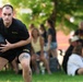 A U.S. Army soldier wearing black PT (physical training) uniform with 'ARMY' printed on the shorts is holding a medicine ball during what appears to be a fitness event or competition. The soldier has visible tattoos on his leg and is in a crouched athletic stance. A crowd of spectators, including families and children, is visible in the blurred background on a grassy field.