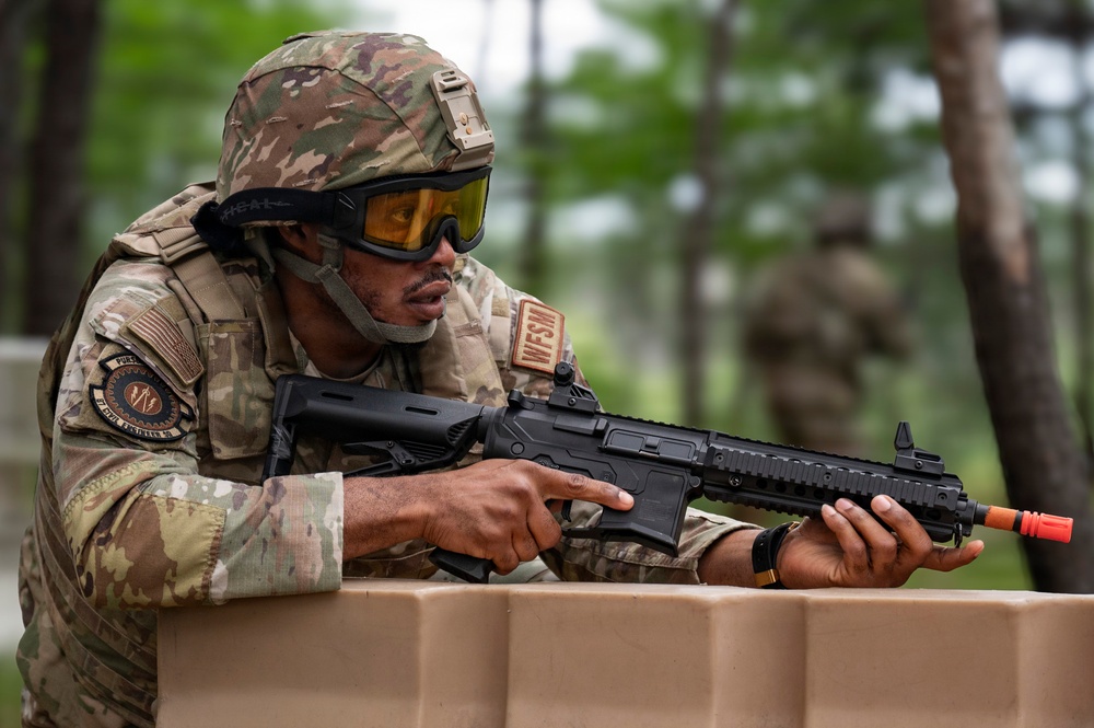A soldier in full multicam camouflage gear is positioned behind a tan barrier, aiming a rifle with an orange muzzle tip indicator. The individual is wearing a tactical helmet with a rail-mounted accessory, amber-tinted protective goggles, and a chest rig with visible unit patches. The setting appears to be an outdoor training exercise in a wooded environment.