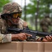 A soldier in full multicam camouflage gear is positioned behind a tan barrier, aiming a rifle with an orange muzzle tip indicator. The individual is wearing a tactical helmet with a rail-mounted accessory, amber-tinted protective goggles, and a chest rig with visible unit patches. The setting appears to be an outdoor training exercise in a wooded environment.