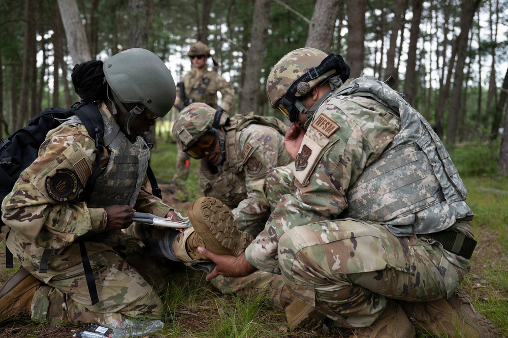 A group of military personnel in camouflage uniforms and tactical gear are conducting what appears to be a field medical training exercise in a forested area. Several soldiers are kneeling on the ground attending to a simulated casualty, with one taking notes on a clipboard while another examines a foot or leg. A fourth service member stands in the background observing the exercise.