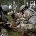 A group of military personnel in camouflage uniforms and tactical gear are conducting what appears to be a field medical training exercise in a forested area. Several soldiers are kneeling on the ground attending to a simulated casualty, with one taking notes on a clipboard while another examines a foot or leg. A fourth service member stands in the background observing the exercise.