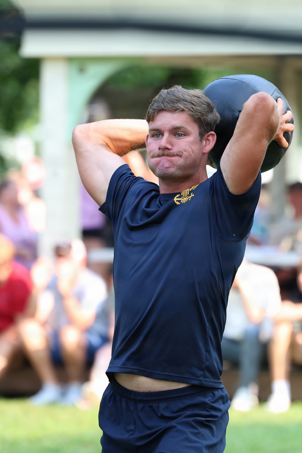A young man with a mustache is holding a dark medicine ball raised above and behind his head, appearing to be in the middle of a throwing or exercise motion. He is wearing a navy blue athletic shirt with a gold emblem on the chest and matching navy shorts. A blurred crowd of spectators is visible in the background, suggesting an outdoor athletic event or competition.