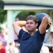 A young man with a mustache is holding a dark medicine ball raised above and behind his head, appearing to be in the middle of a throwing or exercise motion. He is wearing a navy blue athletic shirt with a gold emblem on the chest and matching navy shorts. A blurred crowd of spectators is visible in the background, suggesting an outdoor athletic event or competition.