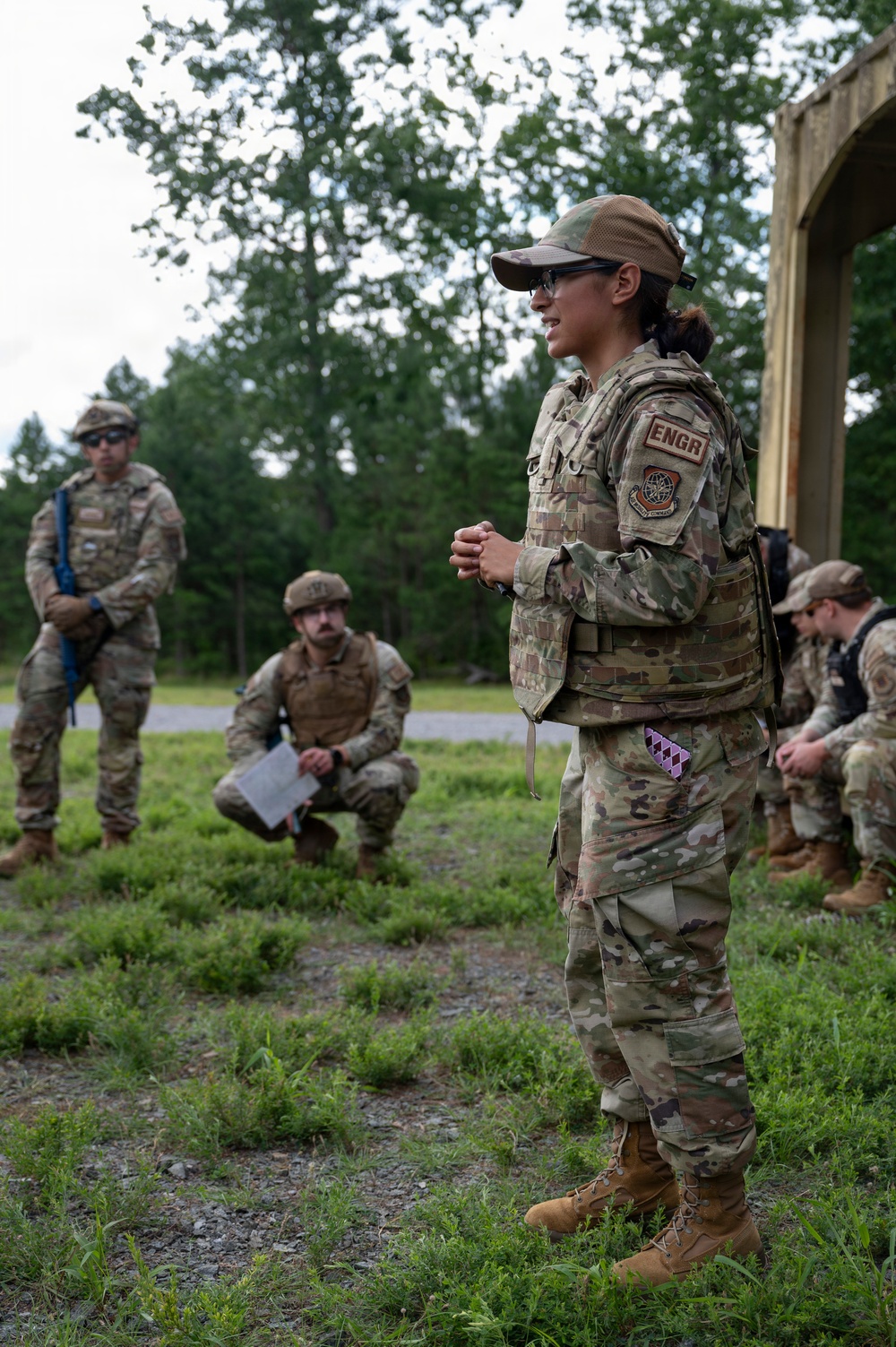 A female military service member wearing OCP (Operational Camouflage Pattern) uniform with an 'ENGR' (Engineer) tab and what appears to be a US Air Force patch is addressing a small group of fellow service members in an outdoor field setting. She appears to be conducting a briefing or instruction session, with personnel listening in various positions around her. The setting suggests a field training exercise or operational planning event.