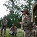 A female military service member wearing OCP (Operational Camouflage Pattern) uniform with an 'ENGR' (Engineer) tab and what appears to be a US Air Force patch is addressing a small group of fellow service members in an outdoor field setting. She appears to be conducting a briefing or instruction session, with personnel listening in various positions around her. The setting suggests a field training exercise or operational planning event.