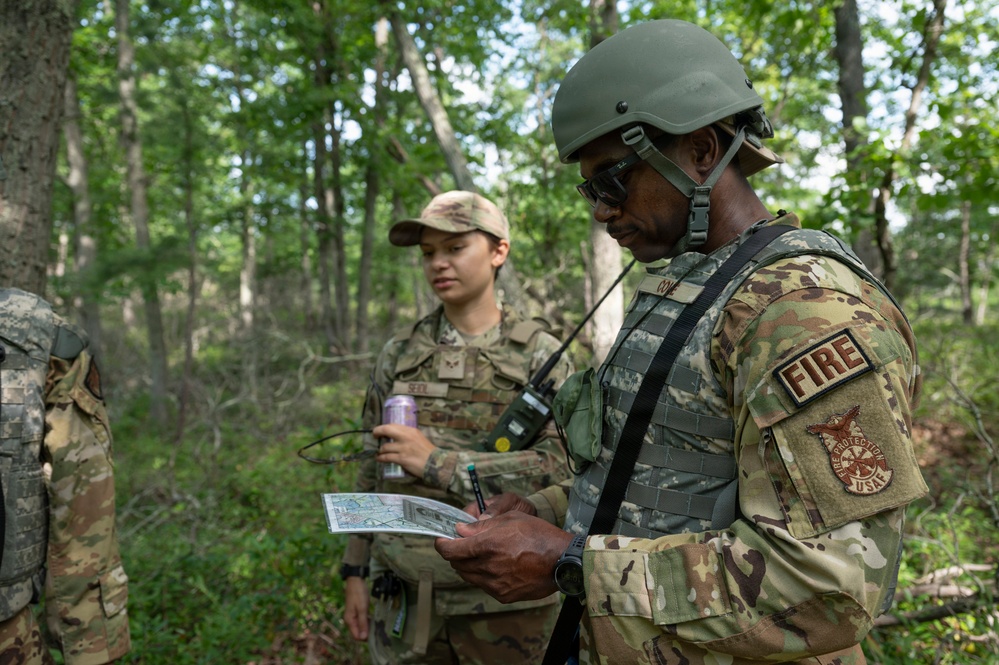 Military personnel in camouflage uniforms are conducting an outdoor field exercise in a wooded area. The foreground figure wears a tactical helmet and body armor with a 'FIRE' patch and a Fire Protection USAF insignia, and is reviewing a document or map. A second service member wearing a patrol cap and holding a radio and a beverage can stands nearby.