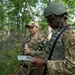 Military personnel in camouflage uniforms are conducting an outdoor field exercise in a wooded area. The foreground figure wears a tactical helmet and body armor with a 'FIRE' patch and a Fire Protection USAF insignia, and is reviewing a document or map. A second service member wearing a patrol cap and holding a radio and a beverage can stands nearby.