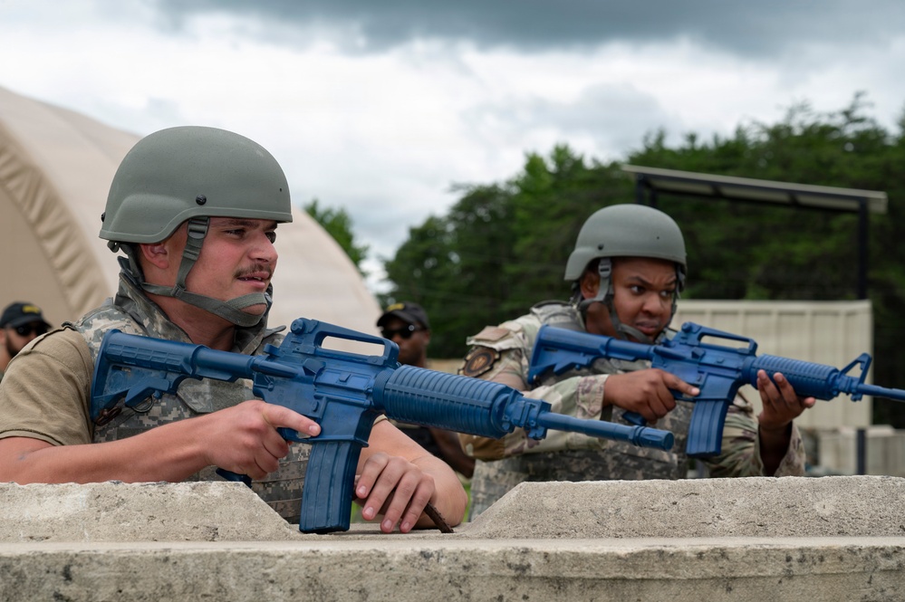 Two military personnel in combat helmets and camouflage uniforms are positioned behind a concrete barrier, holding blue training rifles (inert dummy weapons used for drills). The scene appears to be a military training exercise, indicated by the blue-colored replica firearms which are standard for force-on-force or tactical training scenarios. A tent structure and trees are visible in the background.