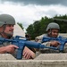 Two military personnel in combat helmets and camouflage uniforms are positioned behind a concrete barrier, holding blue training rifles (inert dummy weapons used for drills). The scene appears to be a military training exercise, indicated by the blue-colored replica firearms which are standard for force-on-force or tactical training scenarios. A tent structure and trees are visible in the background.