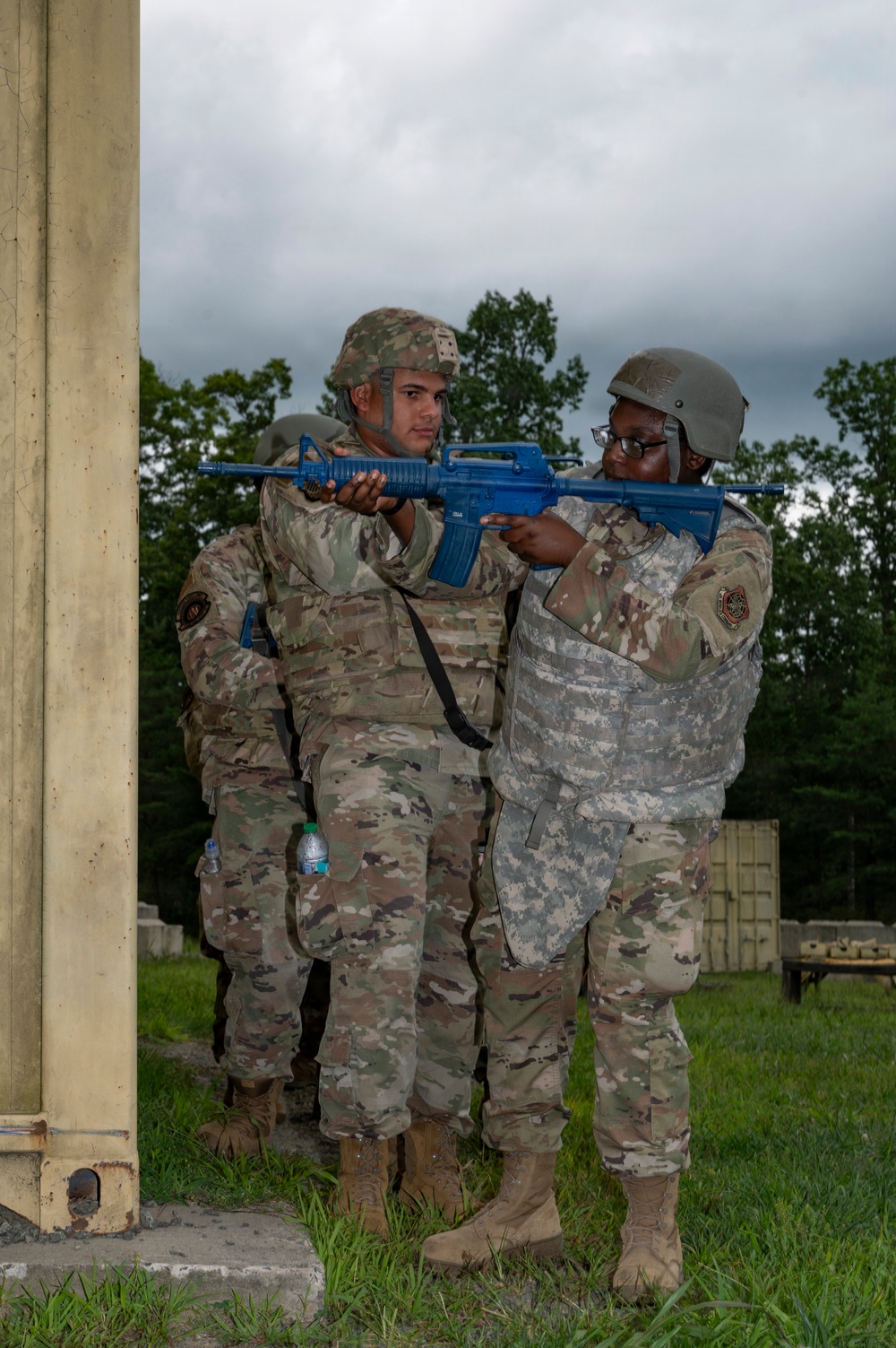 Two military personnel in camouflage uniforms and helmets are engaged in a training exercise near a concrete structure. One soldier wearing older-pattern ACU (Army Combat Uniform) is assisting or instructing another soldier in OCP (Operational Camouflage Pattern) uniform on proper rifle handling technique using blue training rifles. The blue-colored weapons indicate these are inert training/dummy weapons used for tactical drills.