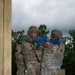 Two military personnel in camouflage uniforms and helmets are engaged in a training exercise near a concrete structure. One soldier wearing older-pattern ACU (Army Combat Uniform) is assisting or instructing another soldier in OCP (Operational Camouflage Pattern) uniform on proper rifle handling technique using blue training rifles. The blue-colored weapons indicate these are inert training/dummy weapons used for tactical drills.