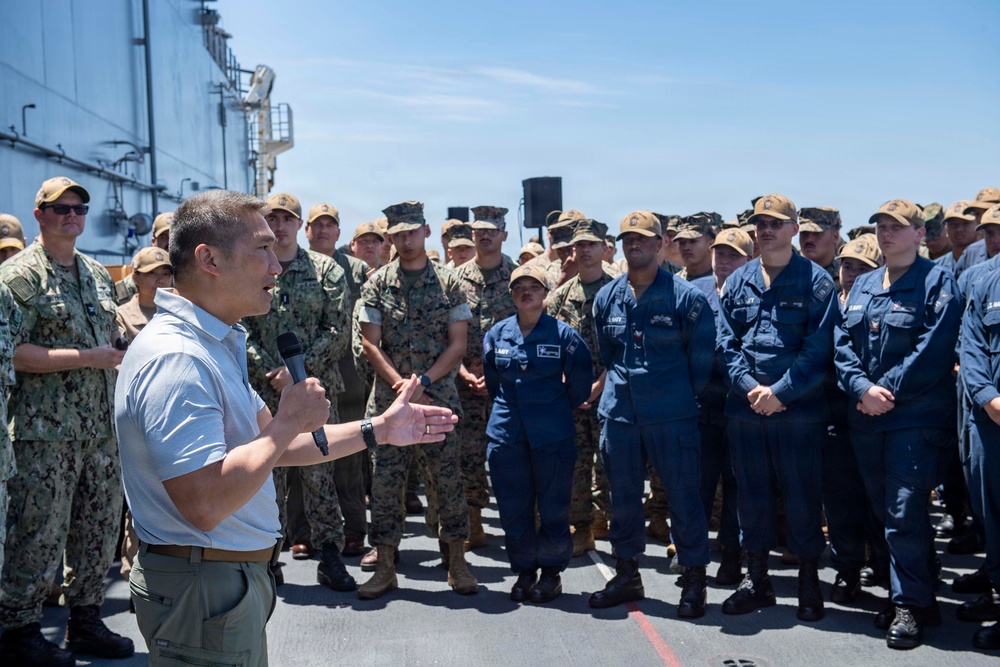 A civilian or senior official in a white polo shirt addresses a large formation of U.S. military personnel on what appears to be the deck of a naval vessel. The audience includes personnel in Navy blue coveralls, Marine Corps MARPAT camouflage uniforms, and Navy NWU Type III camouflage, all wearing tan/coyote brown covers. The setting is an outdoor deck under clear blue skies, suggesting a formal all-hands or command address.