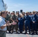 A civilian or senior official in a white polo shirt addresses a large formation of U.S. military personnel on what appears to be the deck of a naval vessel. The audience includes personnel in Navy blue coveralls, Marine Corps MARPAT camouflage uniforms, and Navy NWU Type III camouflage, all wearing tan/coyote brown covers. The setting is an outdoor deck under clear blue skies, suggesting a formal all-hands or command address.