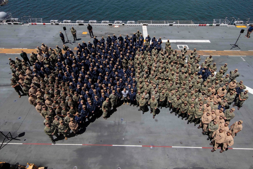 An aerial photograph of a large military group photo taken on the flight deck of a naval vessel, with ocean visible in the background. Personnel are arranged in a deliberate formation wearing multiple uniform types including Navy Working Uniforms (NWU), Marine Corps combat utilities, and dark blue coveralls. The gathering appears to be a formal all-hands or command photo event.