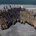 An aerial photograph of a large military group photo taken on the flight deck of a naval vessel, with ocean visible in the background. Personnel are arranged in a deliberate formation wearing multiple uniform types including Navy Working Uniforms (NWU), Marine Corps combat utilities, and dark blue coveralls. The gathering appears to be a formal all-hands or command photo event.