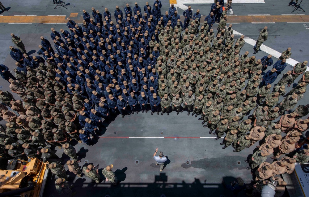 An aerial/overhead photograph shows a large military formation on what appears to be the flight deck of a naval vessel. Personnel are arranged in a semicircular formation, with two distinct uniform groups visible — one in navy blue coveralls and one in camouflage pattern uniforms. A single individual in civilian or light-colored clothing stands at the center base of the formation, appearing to address the assembled personnel.