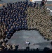 An aerial/overhead photograph shows a large military formation on what appears to be the flight deck of a naval vessel. Personnel are arranged in a semicircular formation, with two distinct uniform groups visible — one in navy blue coveralls and one in camouflage pattern uniforms. A single individual in civilian or light-colored clothing stands at the center base of the formation, appearing to address the assembled personnel.