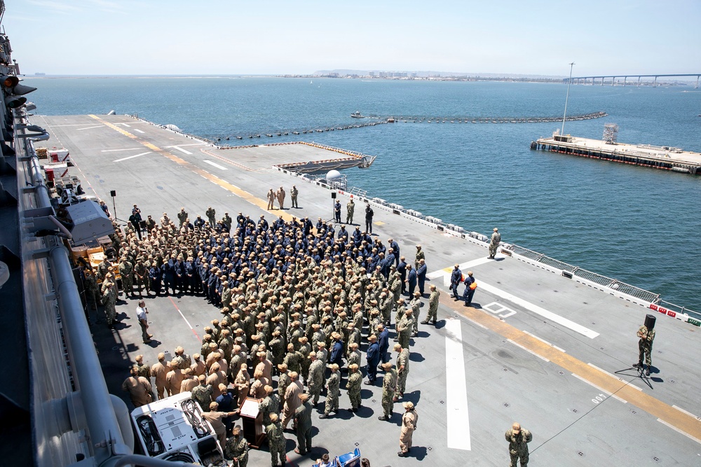 A large military formation ceremony is taking place on the flight deck of a naval vessel, likely an aircraft carrier or amphibious assault ship. Hundreds of uniformed personnel in Navy working uniforms (NWUs) and khaki covers are assembled in formation, with what appears to be a podium or lectern visible, suggesting an official ceremony such as a change of command, commissioning, or award presentation. The ship appears to be docked or anchored in a harbor, with a bridge and coastal infrastructure visible in the background.