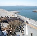 A large military formation ceremony is taking place on the flight deck of a naval vessel, likely an aircraft carrier or amphibious assault ship. Hundreds of uniformed personnel in Navy working uniforms (NWUs) and khaki covers are assembled in formation, with what appears to be a podium or lectern visible, suggesting an official ceremony such as a change of command, commissioning, or award presentation. The ship appears to be docked or anchored in a harbor, with a bridge and coastal infrastructure visible in the background.