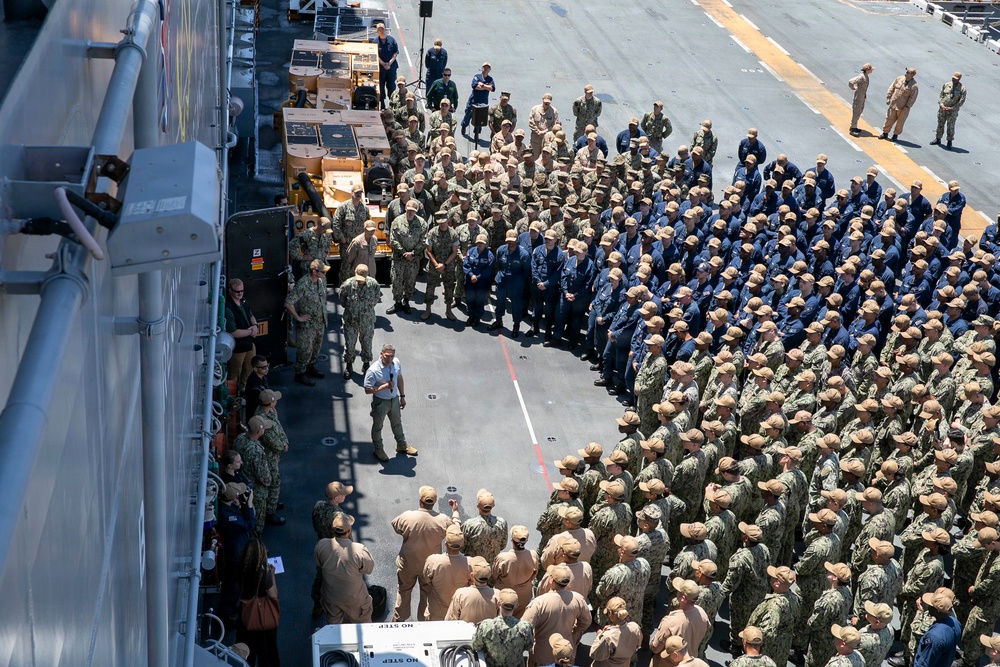 A large military formation is assembled on what appears to be the flight deck of a naval vessel, likely an aircraft carrier or amphibious assault ship. Hundreds of uniformed Navy personnel are arranged in organized rows, with a small group of individuals — including at least one civilian — positioned at the front, suggesting an address, ceremony, or all-hands muster. The image is taken from an elevated angle, revealing the scale of the gathering and surrounding deck equipment.