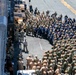 A large military formation is assembled on what appears to be the flight deck of a naval vessel, likely an aircraft carrier or amphibious assault ship. Hundreds of uniformed Navy personnel are arranged in organized rows, with a small group of individuals — including at least one civilian — positioned at the front, suggesting an address, ceremony, or all-hands muster. The image is taken from an elevated angle, revealing the scale of the gathering and surrounding deck equipment.