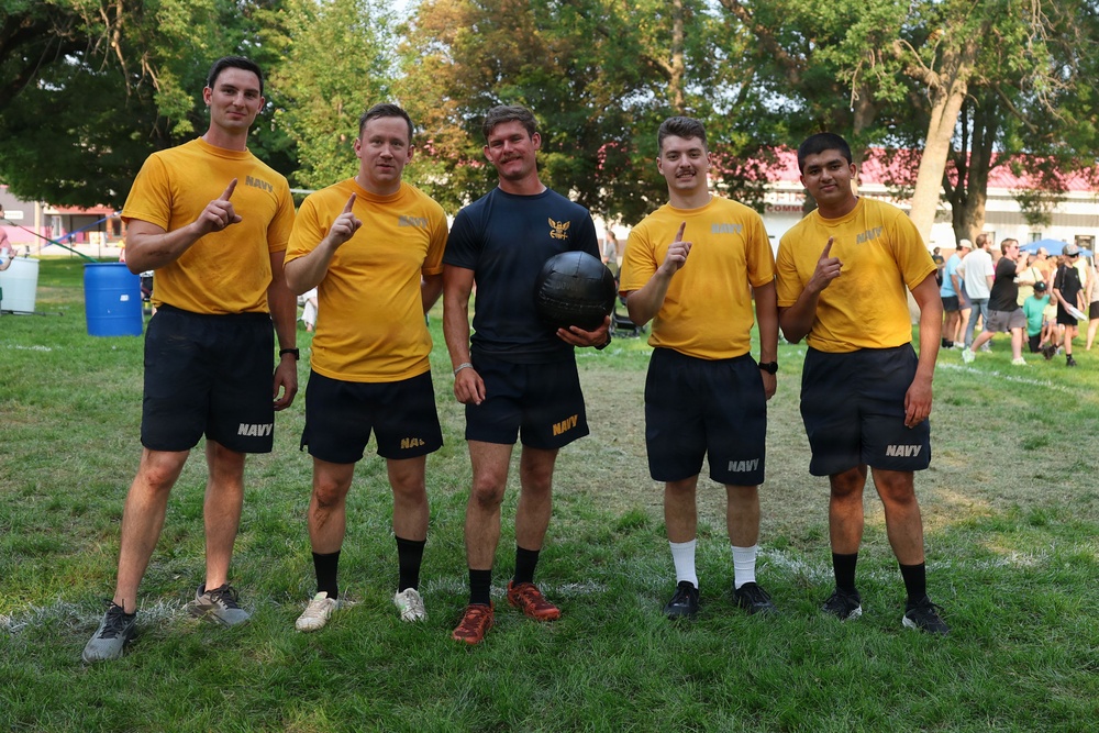 Five individuals wearing U.S. Navy physical training uniforms pose together on a grass field, with four of them holding up index fingers in a 'number one' gesture. The central figure holds a medicine ball or weighted exercise ball and wears a dark navy shirt with a military insignia, suggesting a leadership or instructor role. The setting appears to be an outdoor athletic or recreational event.